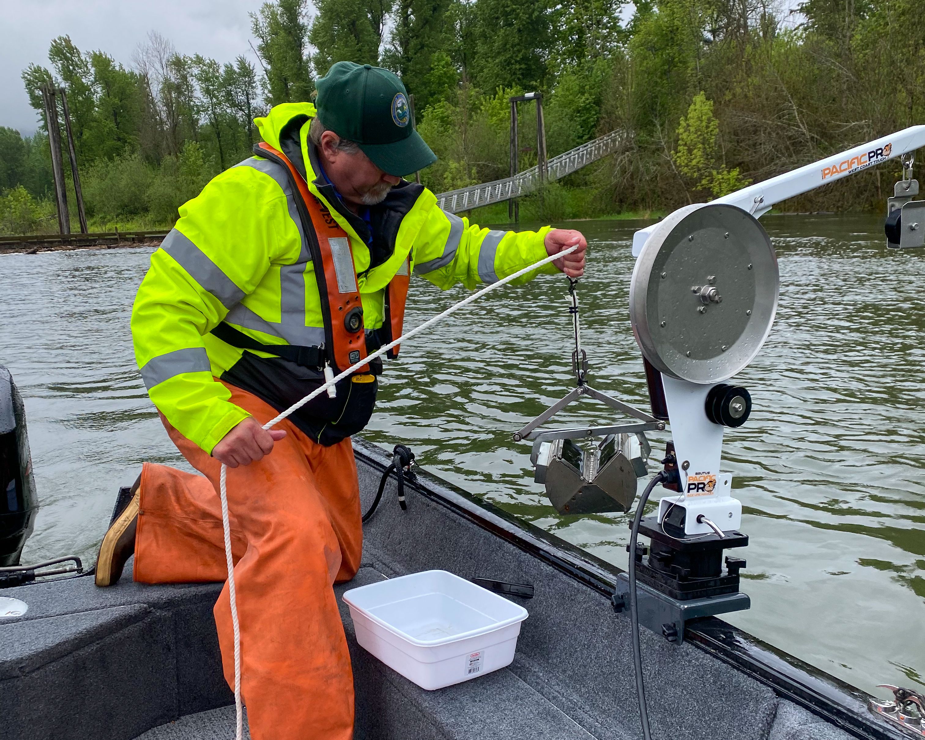 WDFW employee kneeling on a boat wearing bright yellow and orange holding a ponar grab sampler over the edge of the boat.